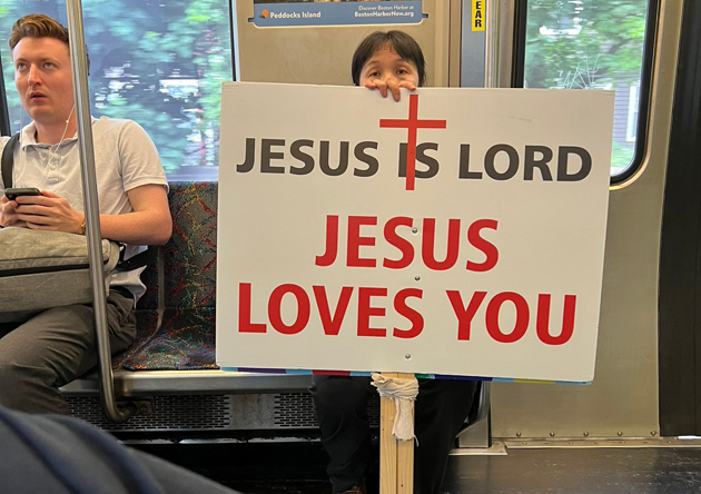Woman riding Red Line from downtown to Braintree with two signs, one reading: Jesus Is Lord, Jesus Loves You